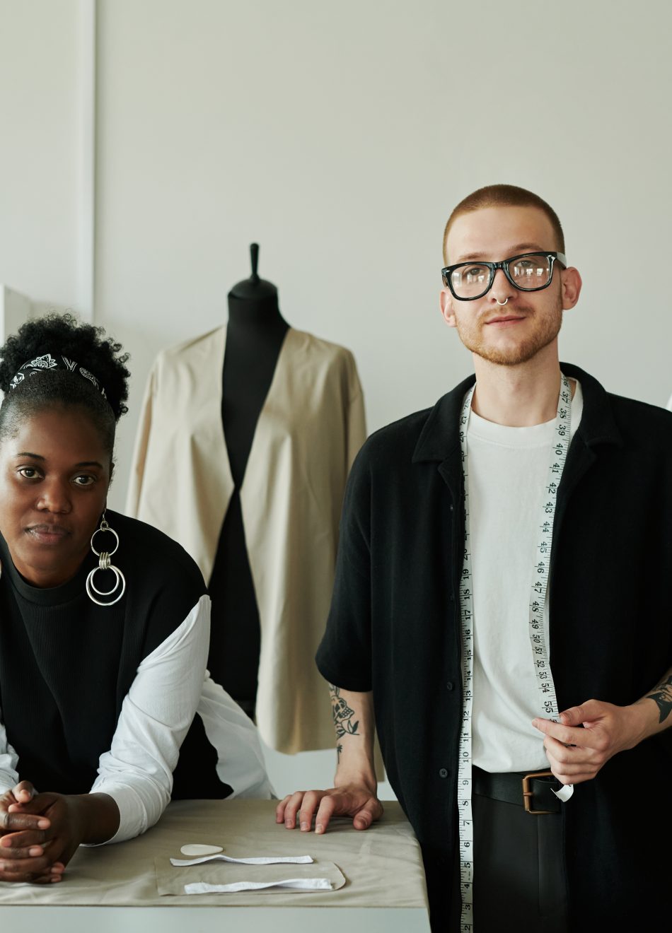 Two young intercultural fashion designers in contrast casualwear looking at camera while standing by workplace with unrolled grey textile